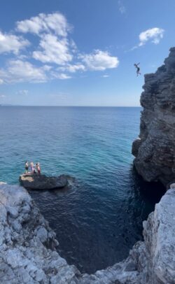 soeone jumping from a cliff into a blue sea, with people stood wathing from a rock in the sea
